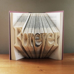 May include: A book with the word "Forever" folded into the pages. The book is open and the pages are folded to create the word in 3D. The book is pink and the pages are white.
