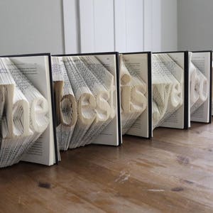 K&ouml;nnte beinhalten: Ein Buchkunstwerk mit den Worten "The best is yet to be", die in die Seiten gefaltet sind. Die B&uuml;cher sind in einer Reihe auf einer Holzoberfl&auml;che angeordnet.