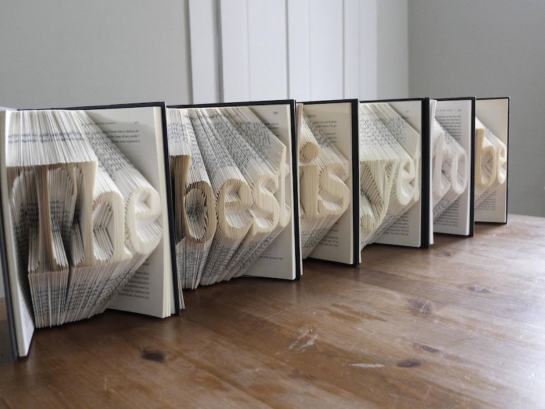 K&ouml;nnte beinhalten: Ein Buchkunstwerk mit den Worten "The best is yet to be", die in die Seiten gefaltet sind. Die B&uuml;cher sind in einer Reihe auf einer Holzoberfl&auml;che angeordnet.