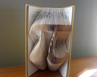 May include: A book folded into the shape of the number 37. The book has a yellow cover and the pages are white. The book is standing on a wooden surface.