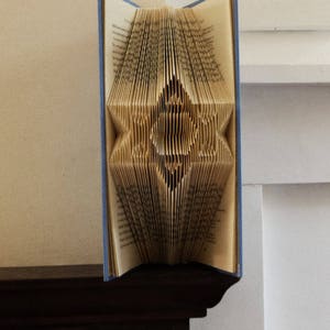 May include: A book folded into a three-dimensional star shape. The book is open and the pages are folded to create a star design. The book is blue and has a white background.