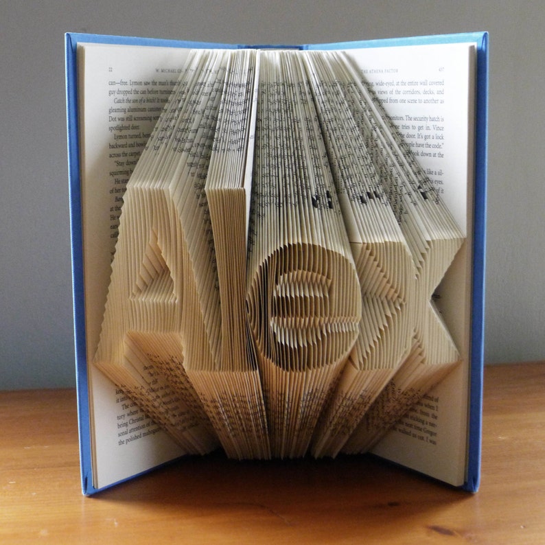 K&ouml;nnte beinhalten: Ein Buch mit dem Namen "ALEX", der in die Seiten gefaltet ist. Das Buch ist blau und die Seiten sind cremefarben.
