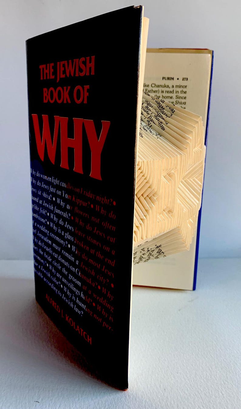 May include: A hardcover book titled "THE JEWISH BOOK OF WHY" with red lettering on a black cover. The pages are folded to create a geometric design. The book is open to a page with text.