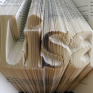 K&ouml;nnte beinhalten: Ein Buch, das mit Hilfe der Seiten in die Form des Wortes &bdquo;LIEBE&ldquo; gefaltet wurde. Das Buch ist ge&ouml;ffnet und die Seiten sind gefaltet, um die Buchstaben zu bilden. Das Buch besteht aus wei&szlig;em Papier und hat einen cremefarbenen Einband.