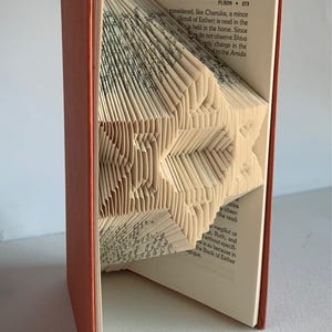 May include: A book with pages folded into a 3D star shape. The book is open and the star is visible. The book cover is orange. The text on the page reads "PURIM * 273 considered, like Chanuka, a minor Scroll of Esther) is read in the is held in the home. Since do not observe Shiva only change in the d to the Amida"