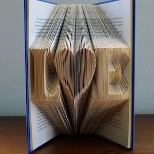 以下が含まれることがあります： 青い表紙の本に「LOVE」の文字が折り込まれています。本が開かれており、ページが折り畳まれて文字が作られています。文字はハートの形をしています。