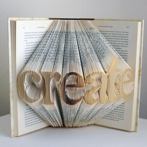 May include: A book sculpture with the word "create" folded into the pages. The pages are fanned out to form the letters, with the text of the book visible. The book is open, and the sculpture is centered.