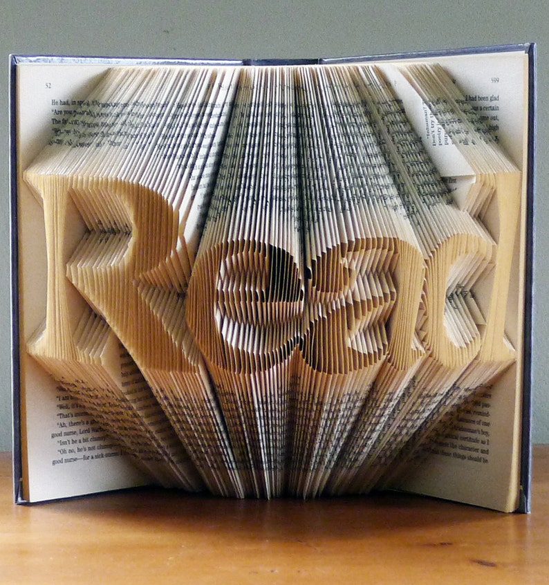 Puede incluir: Un libro con la palabra "READ" doblada en las p&aacute;ginas. El libro est&aacute; abierto y las p&aacute;ginas est&aacute;n dobladas para crear las letras. El libro est&aacute; sobre una superficie de madera.
