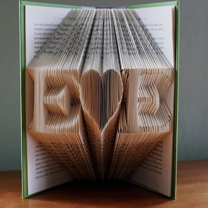 以下が含まれることがあります： ページが「LOVE」という単語の形に折り畳まれ、中央にハートが描かれた本。本が開かれており、ページが折り畳まれて3D効果を生み出しています。
