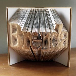 K&ouml;nnte beinhalten: Ein Buch, das in die Form des Wortes "Brenda" gefaltet ist. Das Buch besteht aus wei&szlig;em Papier mit schwarzem Text und ist zu einer 3D-Form gefaltet.