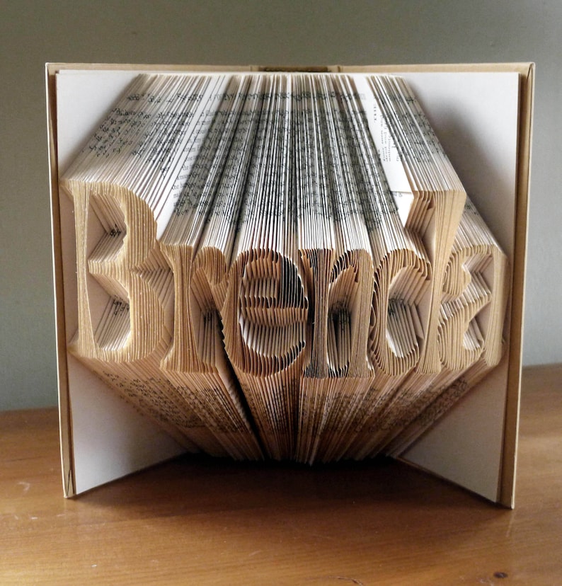 K&ouml;nnte beinhalten: Ein Buch, das in die Form des Wortes "Brenda" gefaltet ist. Das Buch besteht aus wei&szlig;em Papier mit schwarzem Text und ist zu einer 3D-Form gefaltet.