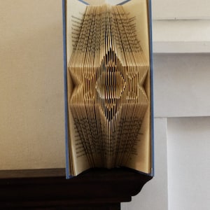May include: A book with blue covers and folded pages, creating a star-shaped design in the center. The pages are beige, and the book is standing upright on a dark wooden surface.