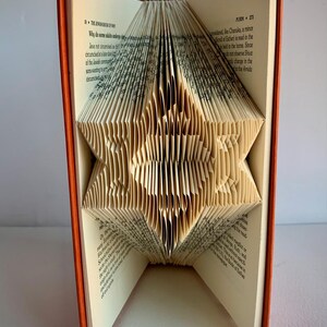 May include: A book with pages folded into a three-dimensional star shape. The book has an orange cover and the pages are cream colored.