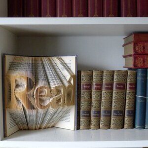 Puede incluir: Un libro doblado en forma de la palabra "READ" en una estanter&iacute;a blanca con otros libros. Los libros de la estanter&iacute;a son principalmente rojos y marrones con letras doradas.