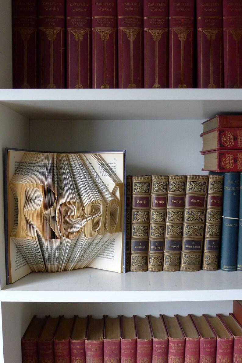 Puede incluir: Un libro doblado en forma de la palabra "READ" en una estanter&iacute;a blanca con otros libros. Los libros de la estanter&iacute;a son principalmente rojos y marrones con letras doradas.
