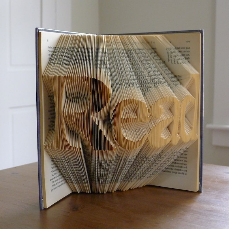 Puede incluir: Una escultura de arte de libro con la palabra "Read" formada doblando las p&aacute;ginas de un libro. El libro est&aacute; abierto y las p&aacute;ginas est&aacute;n dobladas en forma de letras.