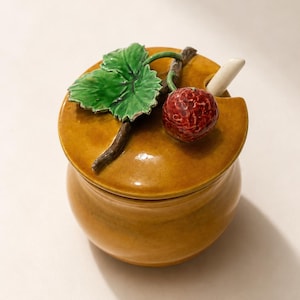 May include: A mustard yellow ceramic jar with a matching lid featuring a sculpted green leaf, a brown stem, and a textured red berry. A small white ceramic spoon is tucked into a notch on the side of the lid, completing this decorative container.
