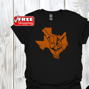 May include: A black short-sleeved t-shirt featuring a distressed orange graphic of the state of Texas with a hand making the hook 'em horns gesture centered on the front. The shirt has a crew neckline and a standard fit.