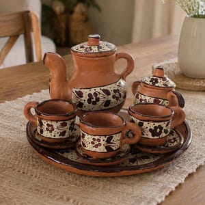 May include: A terracotta tea set featuring a teapot, a sugar bowl, and three cups with matching saucers, all arranged on a round tray. Each piece is decorated with a cream-colored band painted with dark brown floral patterns and small red dots.