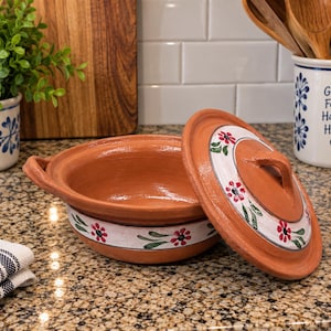 May include: A round terracotta cooking pot with two side handles and a matching lid. The exterior of the pot and the lid feature a white band decorated with hand-painted red flowers and green leaves. The clay has a natural reddish-brown finish.