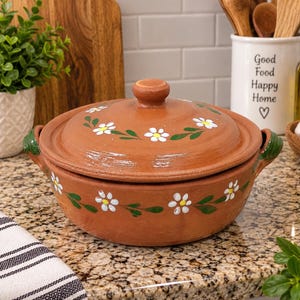 May include: A round terracotta cooking pot with a matching lid, featuring hand-painted white daisy flowers with yellow centers and green leaves. The pot has two green-painted side handles and a central knob on the lid, with a smooth finish.