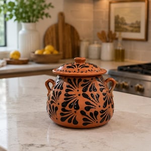 May include: A round terracotta cooking pot with a matching lid, featuring two side handles and a repeating black floral pattern painted across the orange-brown clay surface. The pot has a wide body and a smooth, rounded finish on the exterior.