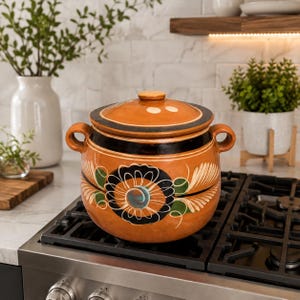 May include: A round terracotta cooking pot with a matching lid, featuring two side handles and a black painted band around the top. The body is decorated with a hand-painted floral motif in black, white, and green, centered on the front surface.