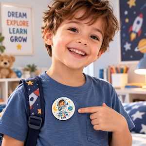 Puede incluir: Un pin circular blanco con un personaje de astronauta de dibujos animados en traje espacial, rodeado de estrellas, una luna y un planeta, con el texto "DREAM BIG" impreso en letras azules y naranjas, sujeto a una camiseta azul.