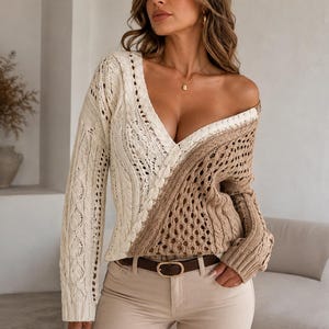 May include: A long-sleeve V-neck sweater featuring a two-tone design with an off-white side and a tan side. The knit garment displays a combination of cable knit patterns and openwork crochet detailing, paired with beige trousers and a brown belt.
