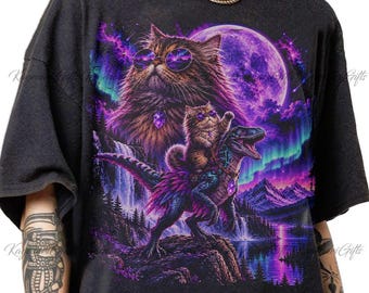 May include: A black t-shirt featuring a vibrant graphic print of a large fluffy cat wearing sunglasses and a crystal pendant, with a smaller cat riding a dinosaur against a backdrop of a purple moon, mountains, a waterfall, and northern lights.