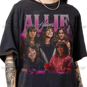 Może przedstawiać: Ciemnoszary t-shirt z krótkim rękawem z nadrukiem graficznym Allie Hayes oraz napisem Allie Hayes dużymi różowymi literami u góry. Projekt zawiera cztery różne portrety fotograficzne tej samej osoby w różnych pozach.