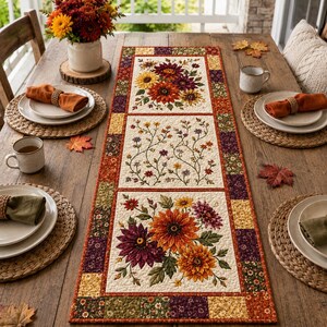 Könnte beinhalten: Ein rechteckiger, gesteppter Tischläufer mit einem Patchwork-Rand in Herbstfarben und drei zentralen Feldern, die florale Drucke von Sonnenblumen, Dahlien und zarten Ranken in Orange-, Gelb-, Rot- und tiefen Violetttönen zeigen.