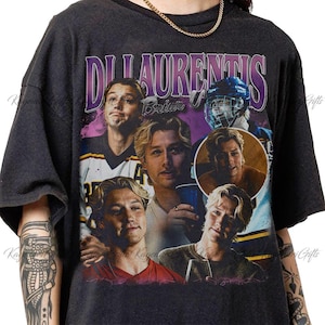 May include: A charcoal gray short-sleeve t-shirt featuring a graphic collage of a person with blonde hair in various poses and settings, including a hockey player. The text "DI LAURENTIS" appears in large purple letters at the top of the design.