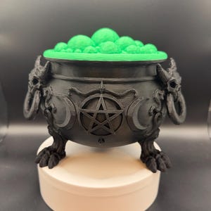 May include: A black cauldron with a matte finish featuring a central pentagram emblem flanked by crescent moons. The vessel has horned skull handles on each side and stands on three clawed feet. It is filled with bright green bubbling liquid.