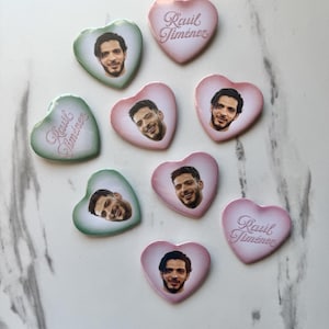 May include: A collection of heart-shaped pins featuring the face of Raul Jiménez or the text Raul Jiménez in cursive script. The pins are colored in shades of soft pink and light green with a glossy finish, arranged in a scattered pattern.