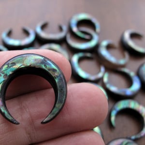 May include: A collection of crescent moon shaped earrings made from abalone shell. The earrings have a black base and iridescent green, blue, and gold accents.