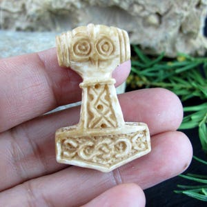 Può includere: Un ciondolo color crema a forma di martello di Thor, Mjolnir. Il martello presenta intagli intricati, tra cui motivi geometrici e disegni a spirale. Il ciondolo è tenuto in mano, mostrando la sua dettagliata maestria e il tema vichingo.