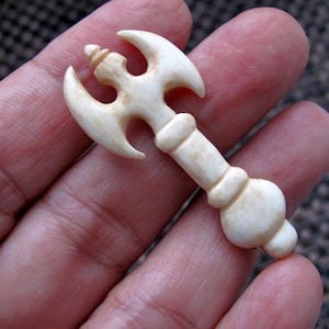 以下が含まれることがあります： オフホワイトの彫刻が施されたミニチュアの斧ペンダント。 斧の頭は湾曲した刃を持ち、柄には丸みを帯びたディテールがあります。 ペンダントは人の手で持たれており、そのサイズと複雑なデザインを示しています。