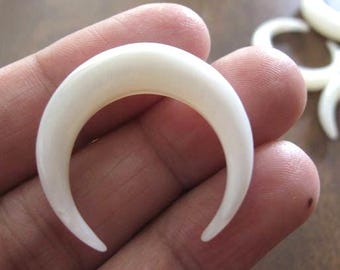 May include: A collection of off-white, crescent-shaped jewelry pieces. The curved, horn-like shapes are smooth and polished, with a slight sheen. The pieces are held in a hand, suggesting they are earrings or pendants.