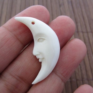 Puede incluir: Un colgante de media luna tallada, de color blanquecino, con un rostro sereno. La luna tiene un pequeño agujero en la punta para ensartar. El colgante se sostiene sobre un fondo borroso.