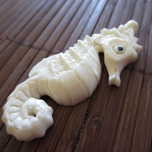 May include: A white carved seahorse figurine with a detailed design. The seahorse is lying on a brown bamboo mat.