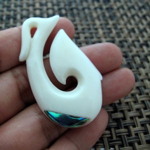 Puede incluir: Pendiente de hueso blanco tallado en forma de anzuelo de pesca tradicional maorí, con una incrustación de abulón verde y azul.