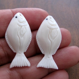 May include: Two white carved bone fish pendants with black eyes. The fish are detailed with carved scales and fins.