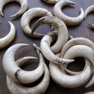 May include: A collection of wooden crescent-shaped earrings in a light brown color with dark brown streaks. The earrings are arranged in a pile on a brown surface.
