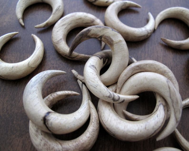 May include: A collection of wooden crescent-shaped earrings in a light brown color with dark brown streaks. The earrings are arranged in a pile on a brown surface.