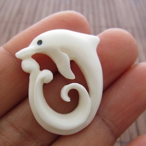 Könnte beinhalten: Weißer, geschnitzter Delfin-Anhänger aus Knochen mit einem schwarzen Auge. Der Delfin ist zu einer Spirale geformt.