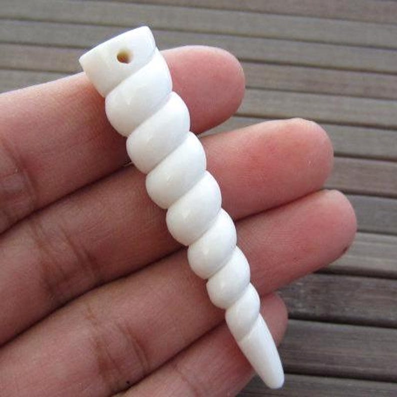 Op de afbeelding: Een witte, spiraalvormige hanger of kraal, die lijkt op een zeeschelp. De kraal heeft een geboord gat aan de bovenkant en een glad, gepolijst oppervlak. Het ontwerp doet denken aan een schelp, perfect voor het maken van sieraden of knutselen.
