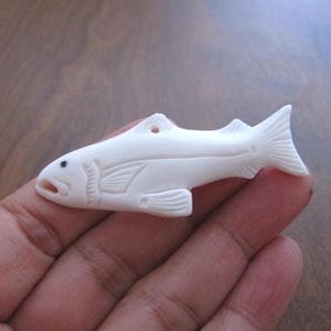May include: A white carved bone fish pendant. The fish is detailed with fins and a mouth. The pendant has a hole at the top for hanging.