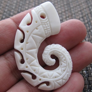 May include: White bone pendant with intricate carvings in a spiral shape. The pendant has a hole at the top for hanging.