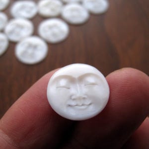 May include: A collection of white, circular beads, each carved with a smiling face. The main bead is held between fingers, showcasing the detailed facial features. The beads have a smooth, polished appearance, suggesting they are made of a stone-like material.